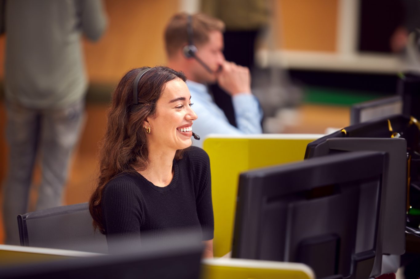 Businesswoman In Cubicle Wearing Headset Talking To Caller In Busy Customer Services Centre